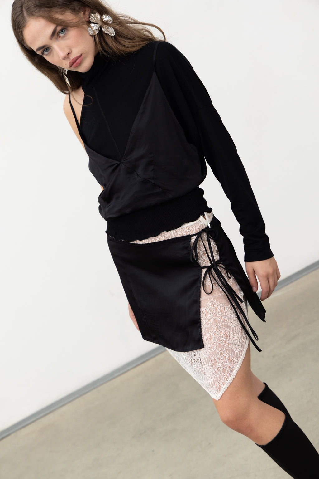 Model wearing the Sacro Top, a black long-sleeve layered top in jersey and satin, styled with a satin and lace wrap mini skirt